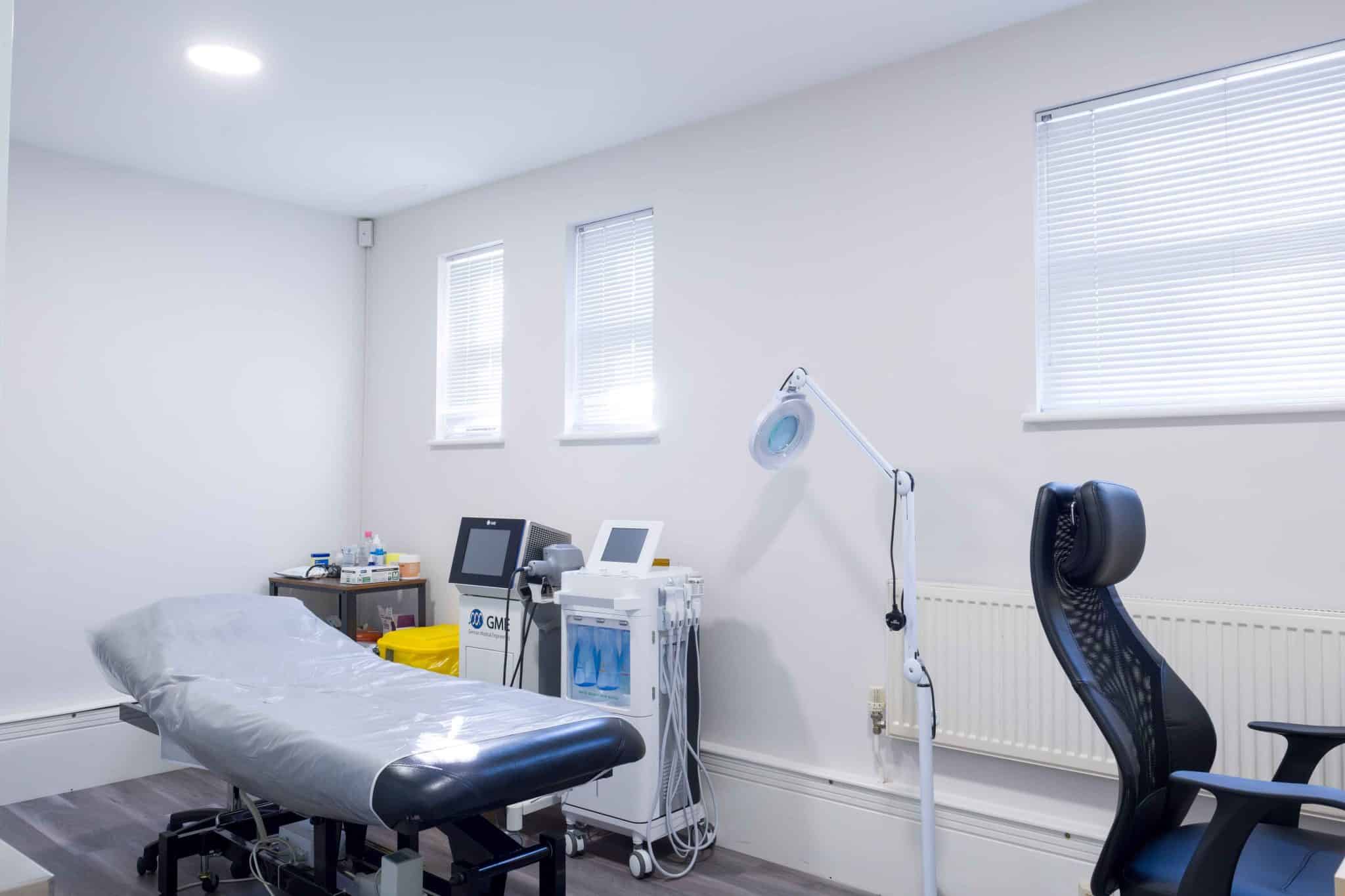 A bright medical examination room with a padded patient bed, monitoring equipment, and a tall lamp near a window with blinds.