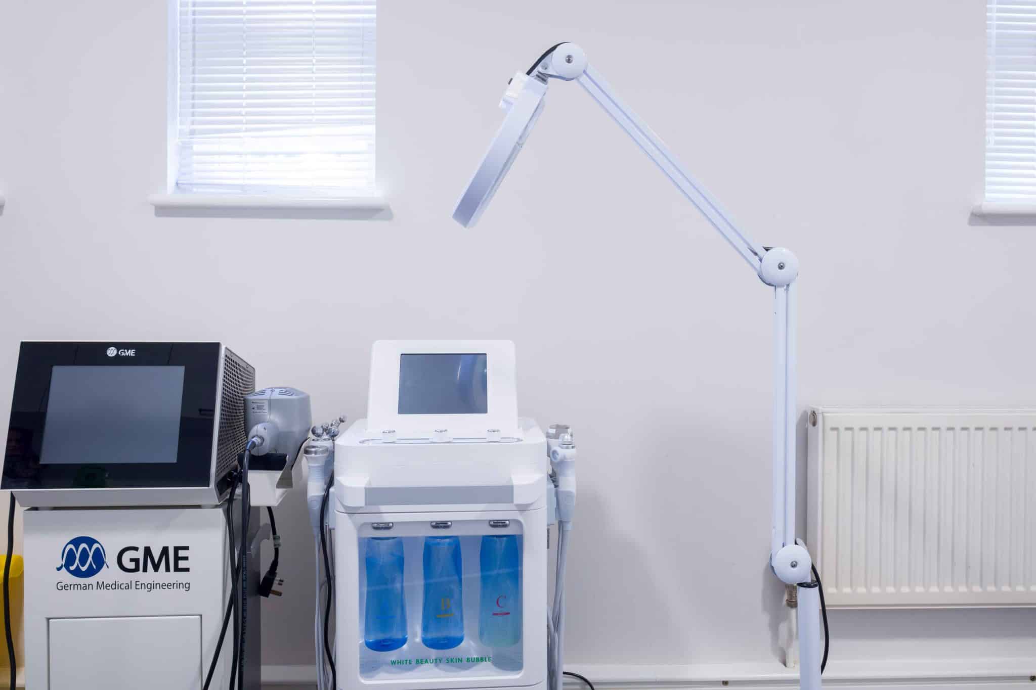 White medical device with touchscreen and a long adjustable lamp in a clinical room; windows with blinds behind.