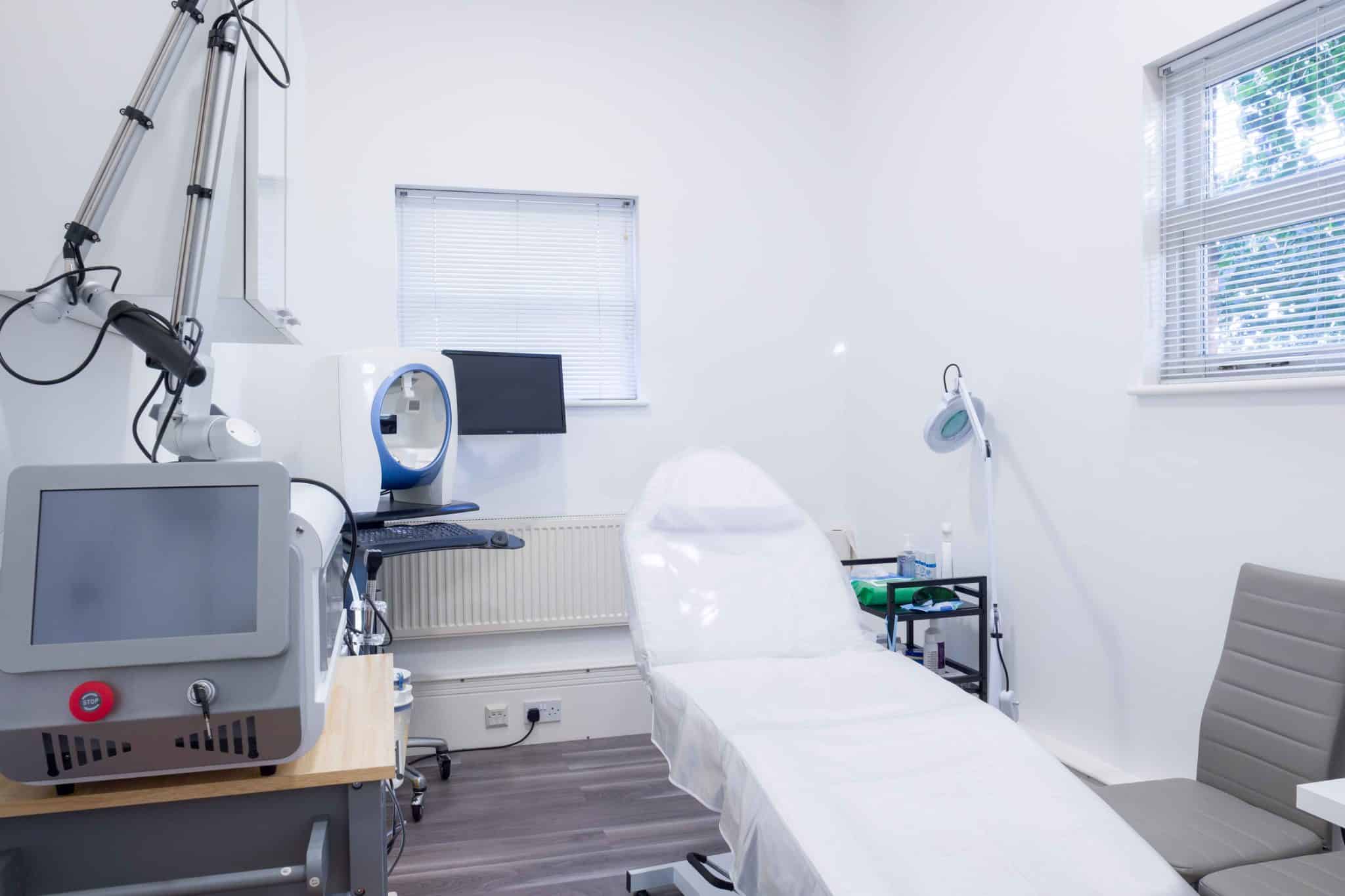 Modern medical examination room with a white recliner chair, monitoring equipment, and a window with blinds in the background.