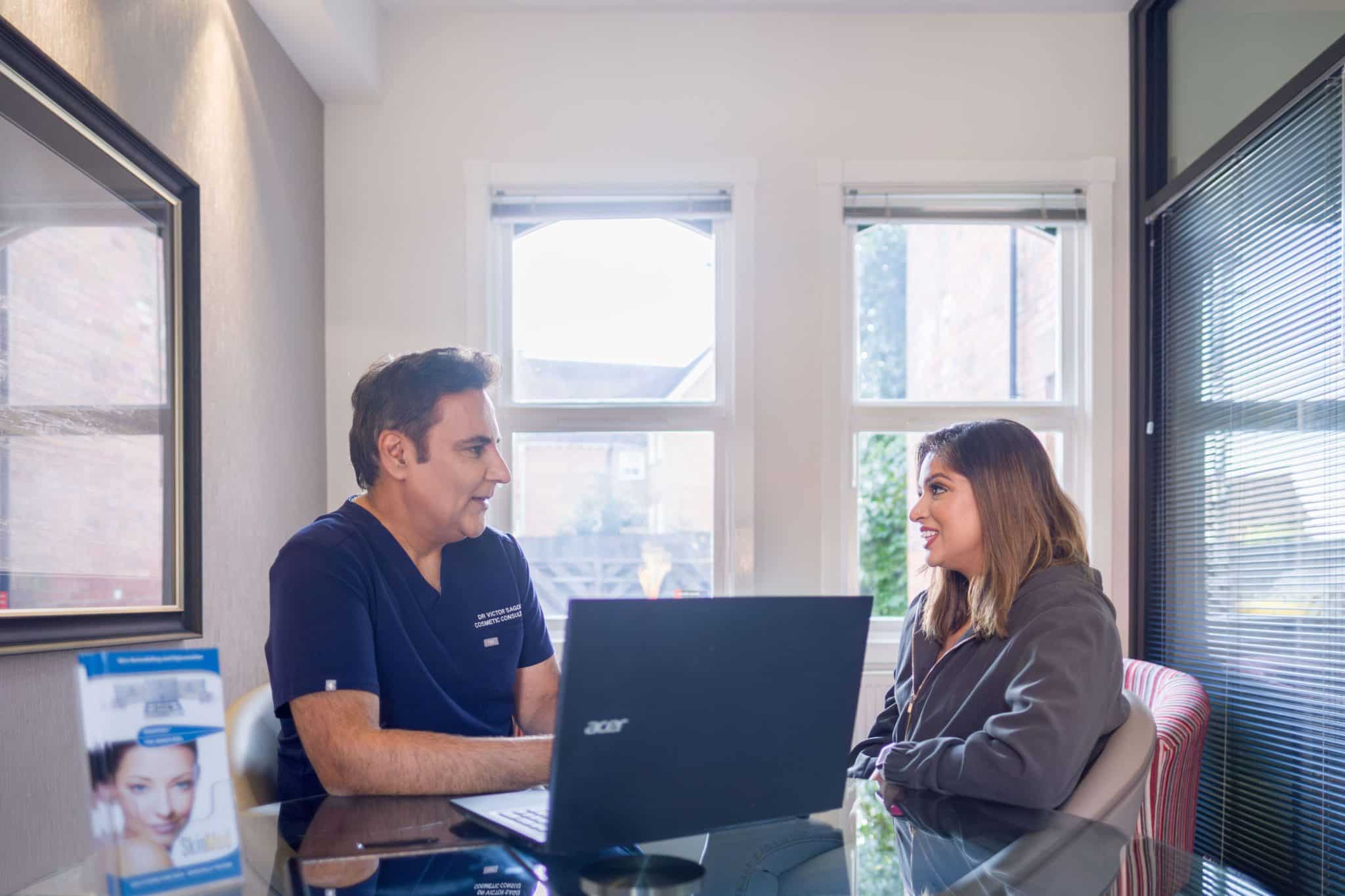 Two colleagues seated at a glass desk in a bright office, talking with a laptop between them.