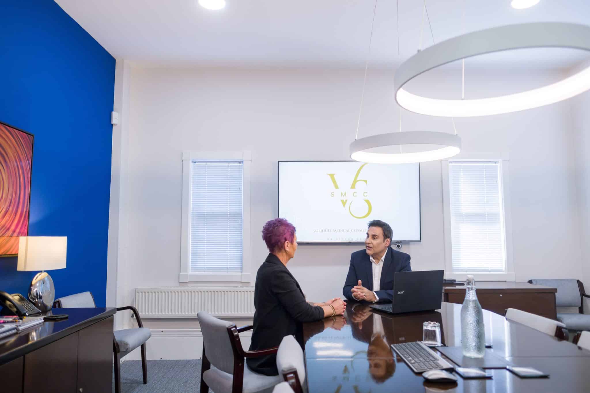 Two professionals sit at a glossy conference table, talking with a presentation screen in the background displaying a logo.