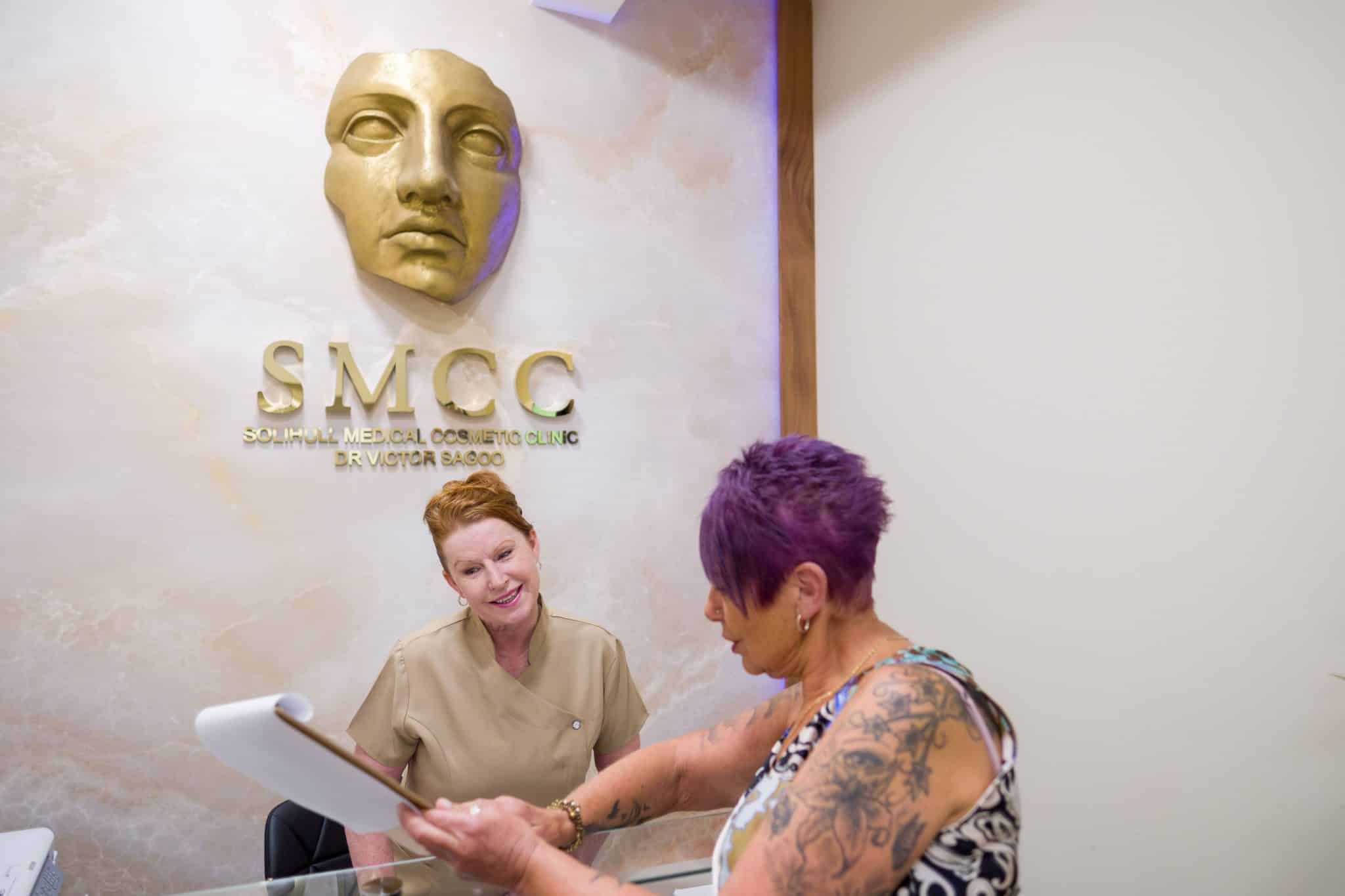 Reception area of a cosmetic clinic with two women reviewing documents at a glass desk; a gold face sculpture and sign appear on the wall behind them.