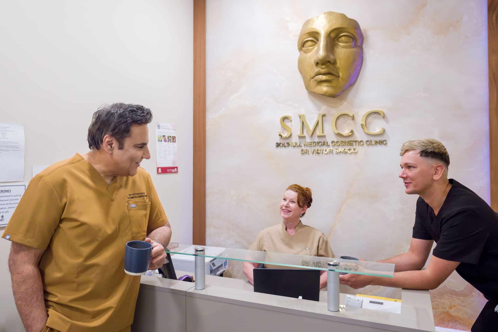 Reception area of a cosmetic clinic with staff in beige scrubs conversing near a glass counter; gold wall sculpture and 'SMCC' sign in the background