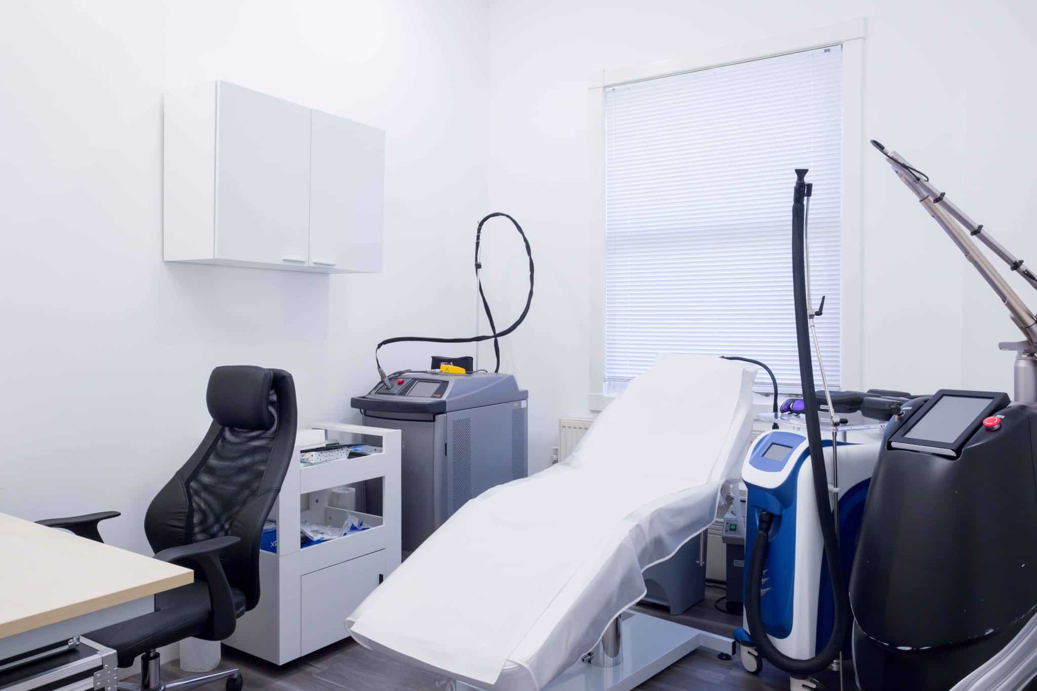 Bright clinic room with a reclining patient chair, white cabinets, and medical equipment by a window with blinds.