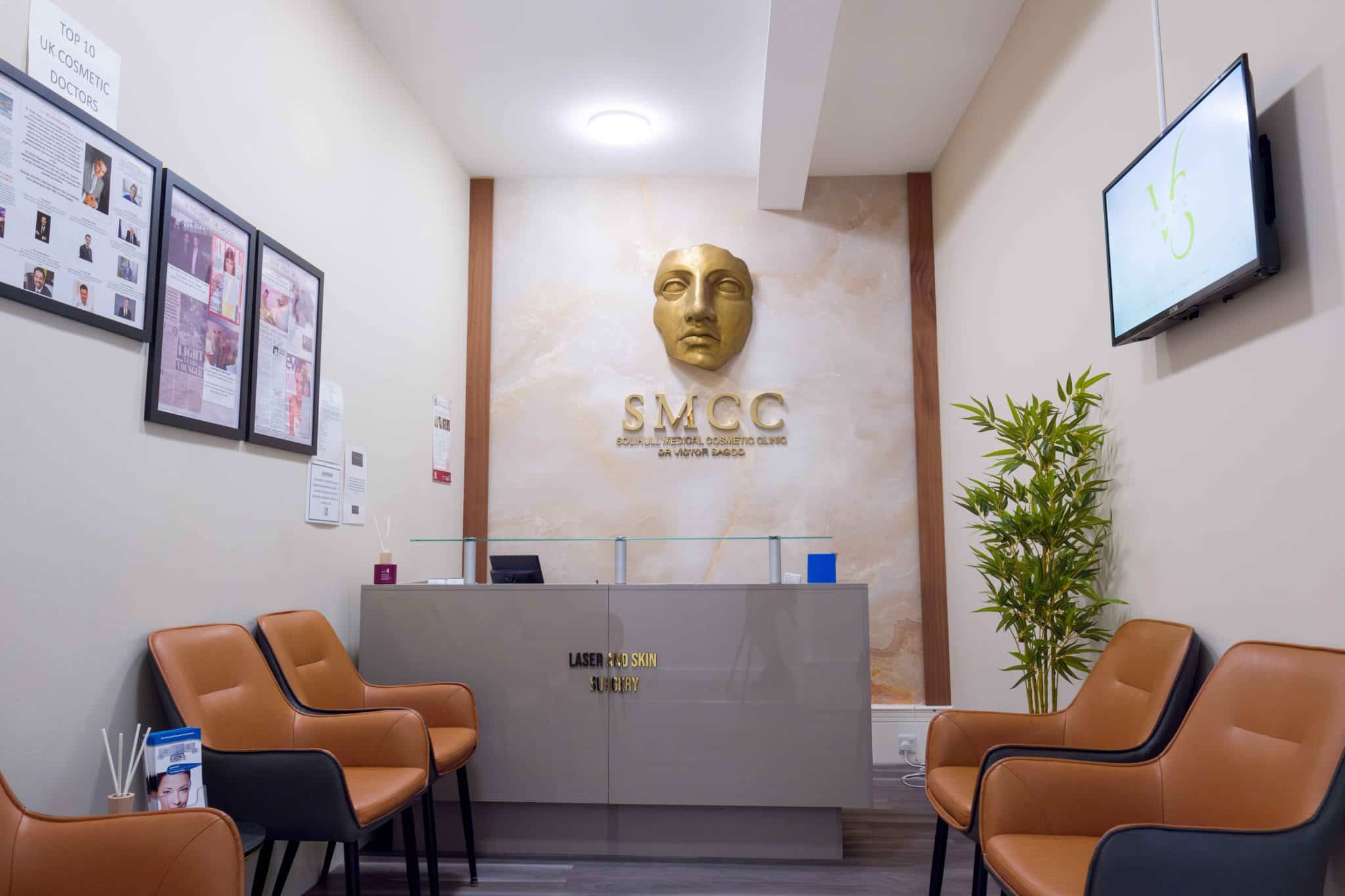Waiting area of a cosmetic clinic reception: beige walls, a gold mask sculpture on the wall behind a gray reception desk, framed posters on the left, a TV on the right, and rows of tan leather chairs.