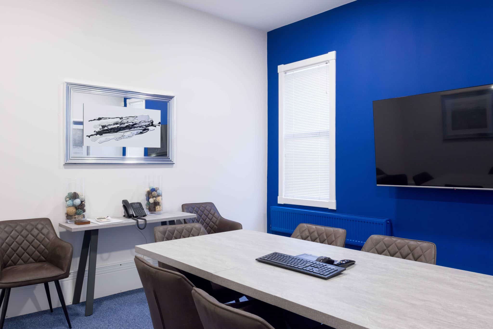 Bright conference room with a blue accent wall, white blinds, and a wall-mounted TV above a radiator; multiple gray tables and brown quilted chairs arranged for a meeting.