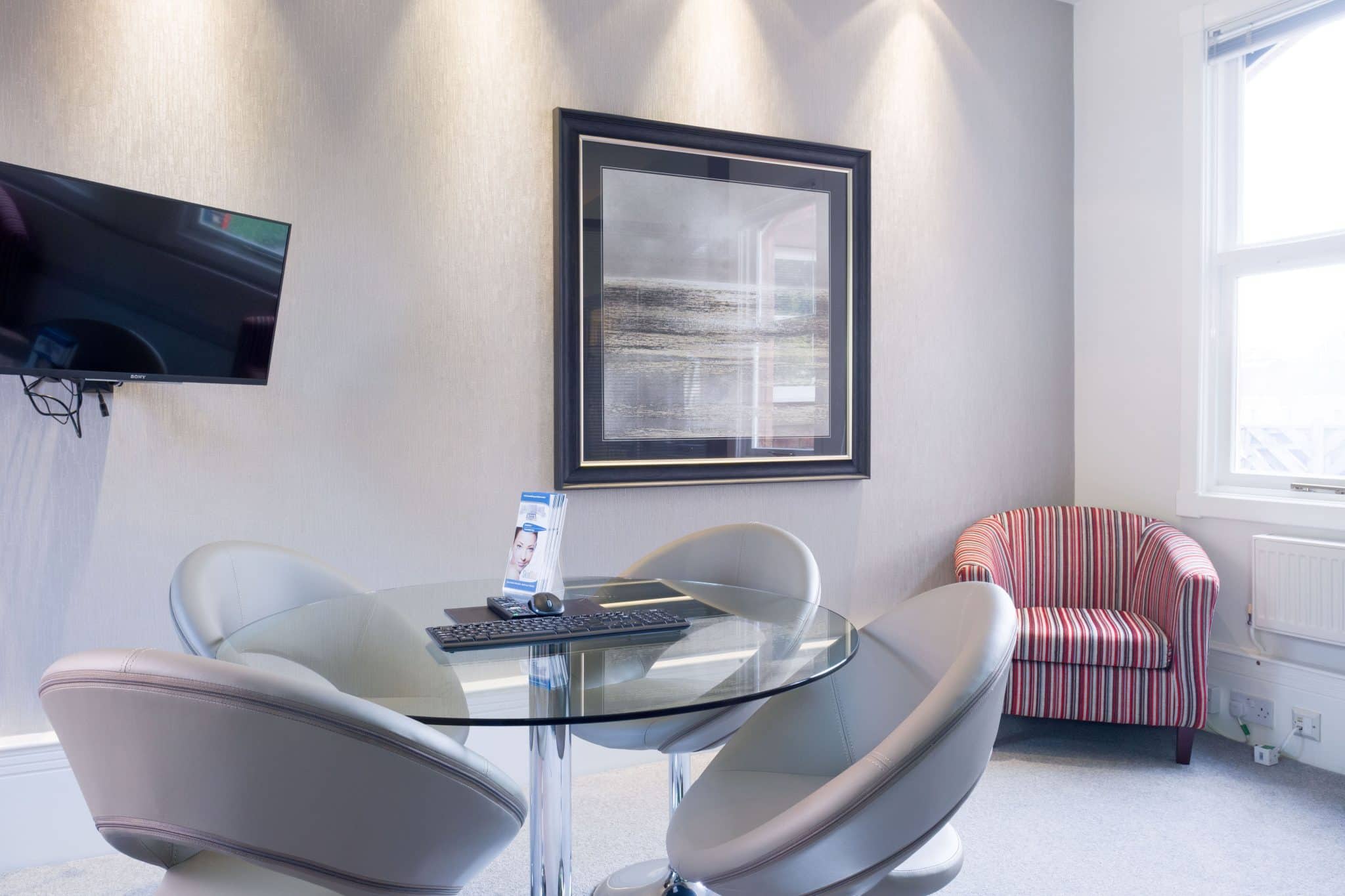 Modern office/lounge with a glass round table, three gray swivel chairs, and a striped red chair beside a large framed seascape on textured wall.