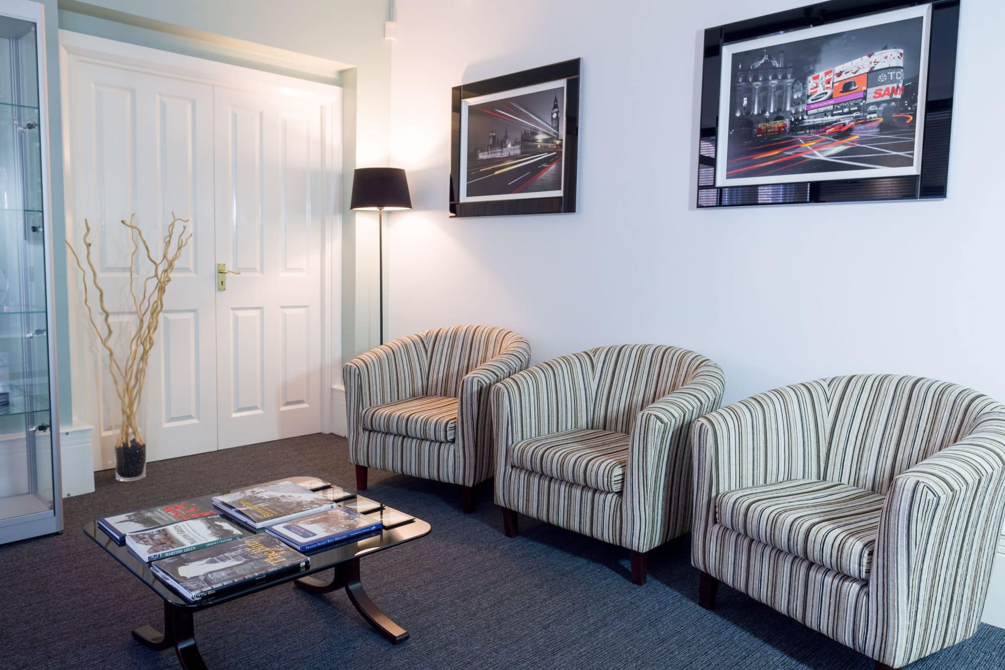 Cozy waiting area with striped upholstered armchairs around a glass coffee table laden with magazines on a dark carpeted floor.
