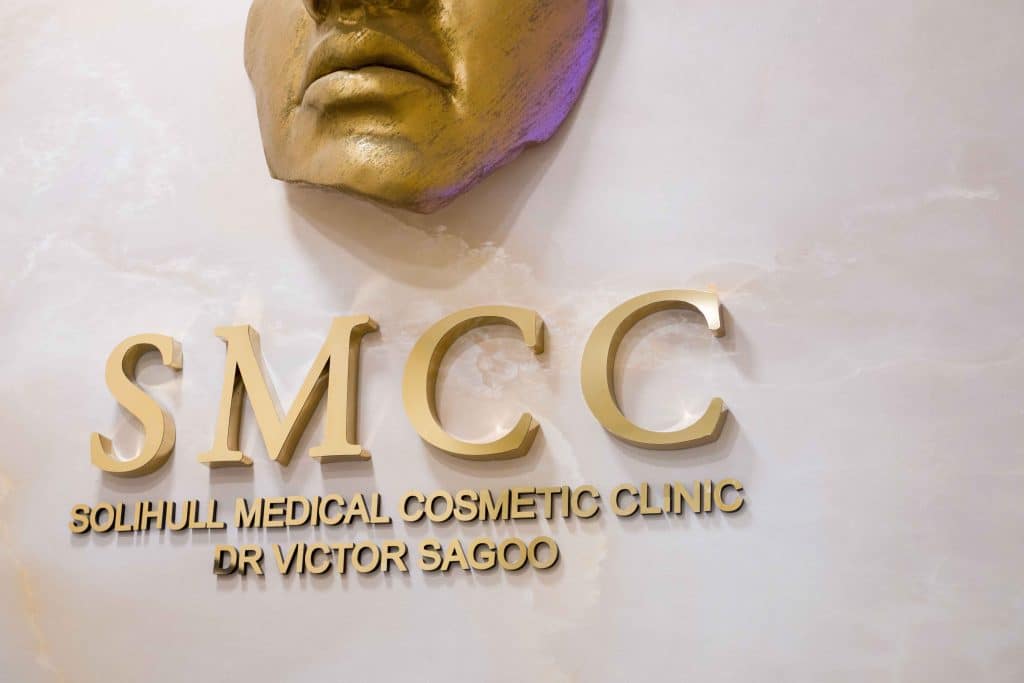 Partial gold face sculpture with large gold letters 'SMC' and the sign reading Solihull Medical Cosmetic Clinic and Dr Victor Sagoo on a pale wall.